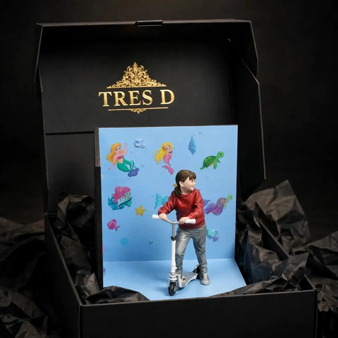 Diorama de niña en patinete dentro de caja premium negra TRES D con logo dorado, embalaje de lujo coleccionista